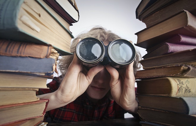 積み上げられた本と本の間から双眼鏡で覗く人